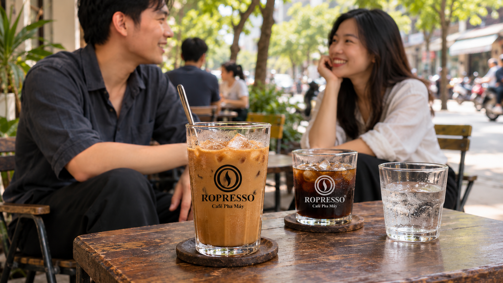 Ly cà phê sữa đá và cà phê đen đá có logo Ropresso trên bàn quán ngoài trời.